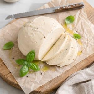 Homemade cashew coconut mozzarella sliced on parchment paper with fresh basil, olive oil, sea salt and black pepper on a wooden board.